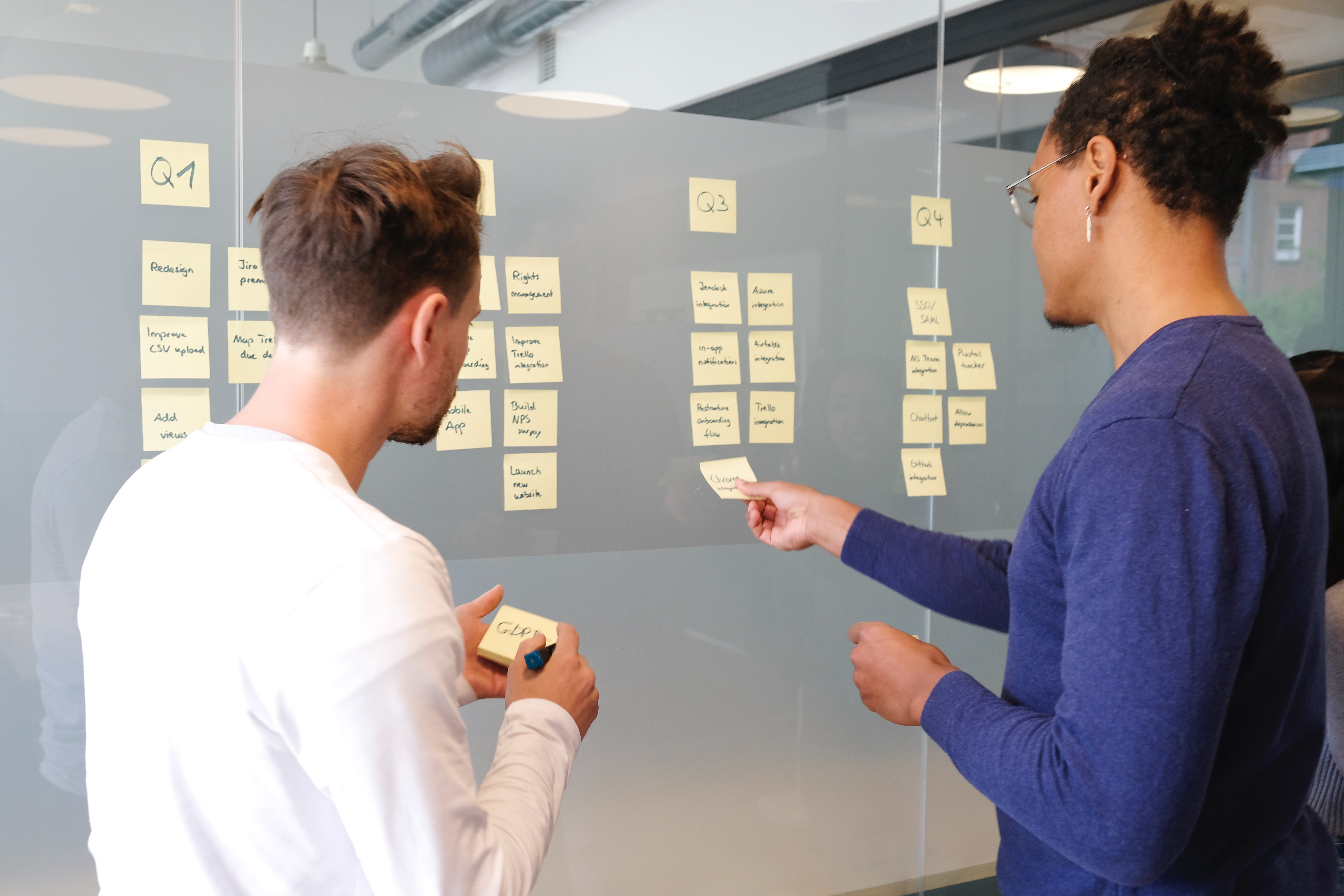 Two individuals in a collaborative planning session using sticky notes on a glass wall to organize tasks by quarters, with one person placing a note.